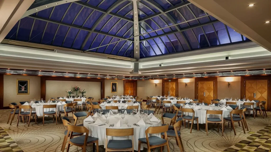 A spacious banquet hall with round tables covered in white tablecloths, neatly set with glassware and napkins, under a large glass ceiling.
